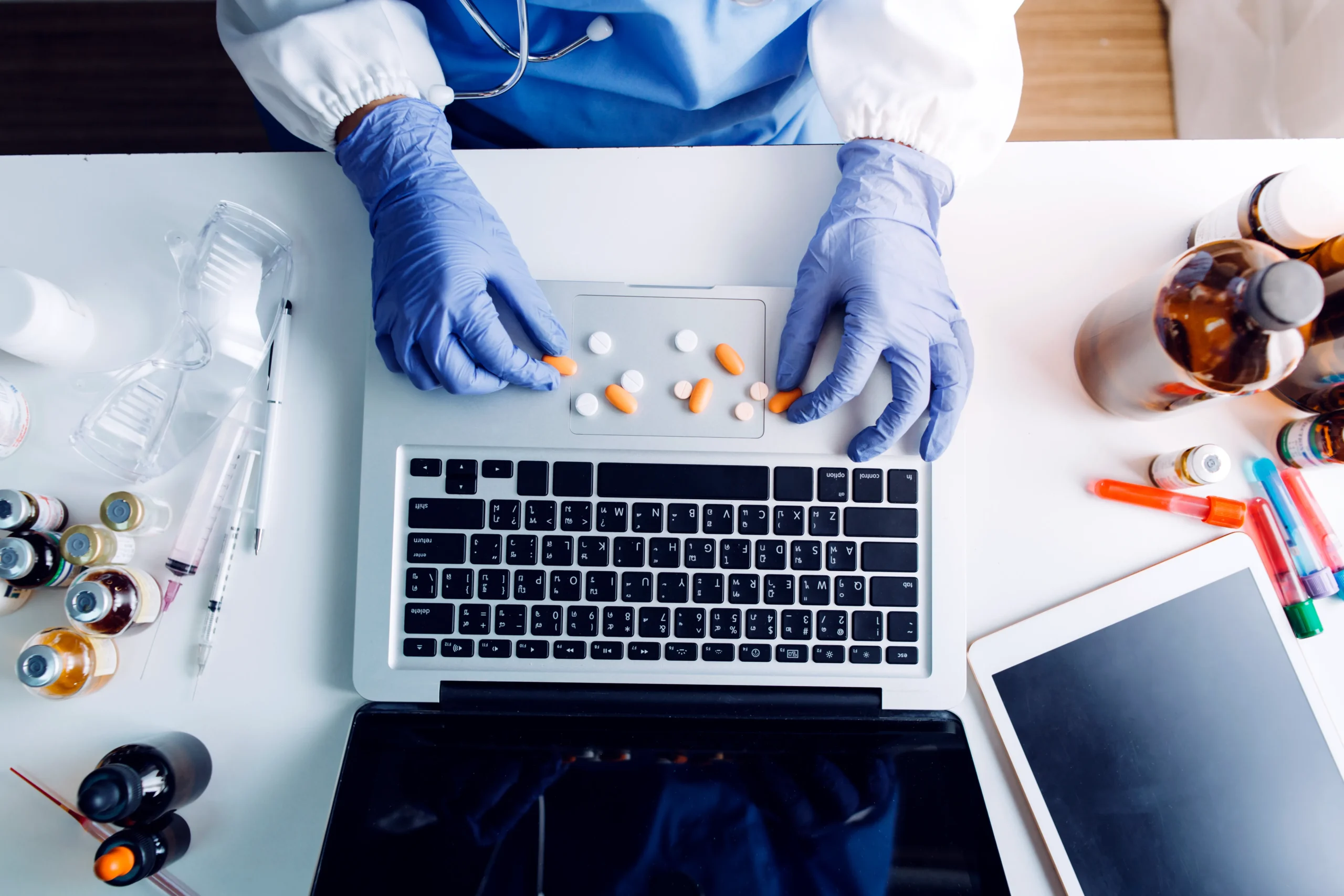 Top-down view of a clinician in blue gloves sorting pills on a laptop desk for Drug Data API management.