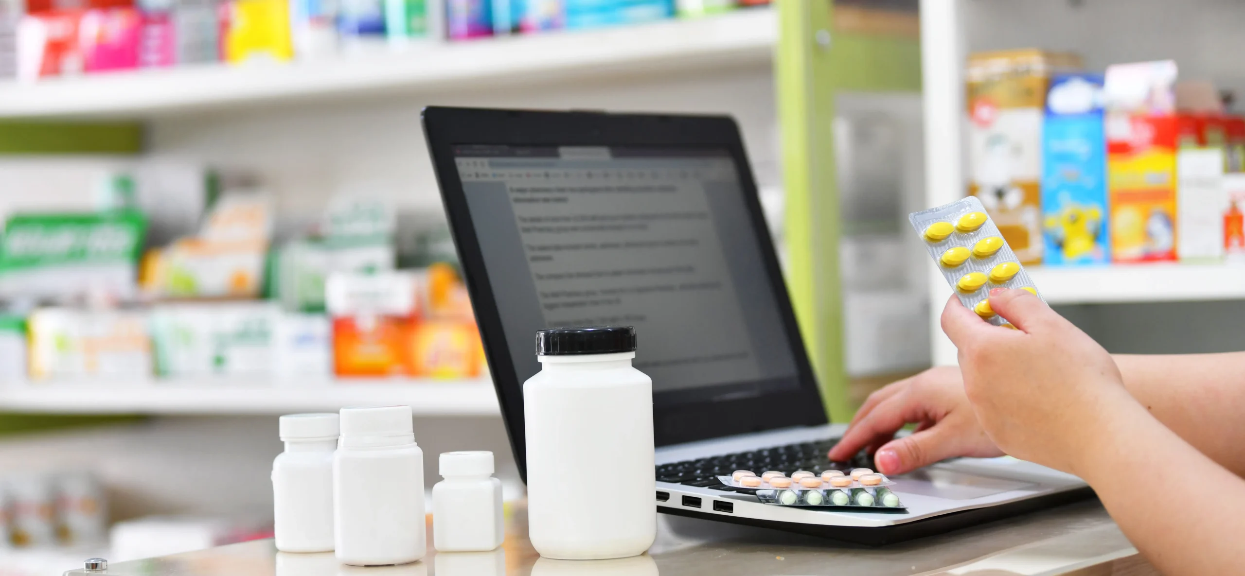 Pharmacist typing on a laptop while inspecting medication, using a drug information API to check prescriptions.